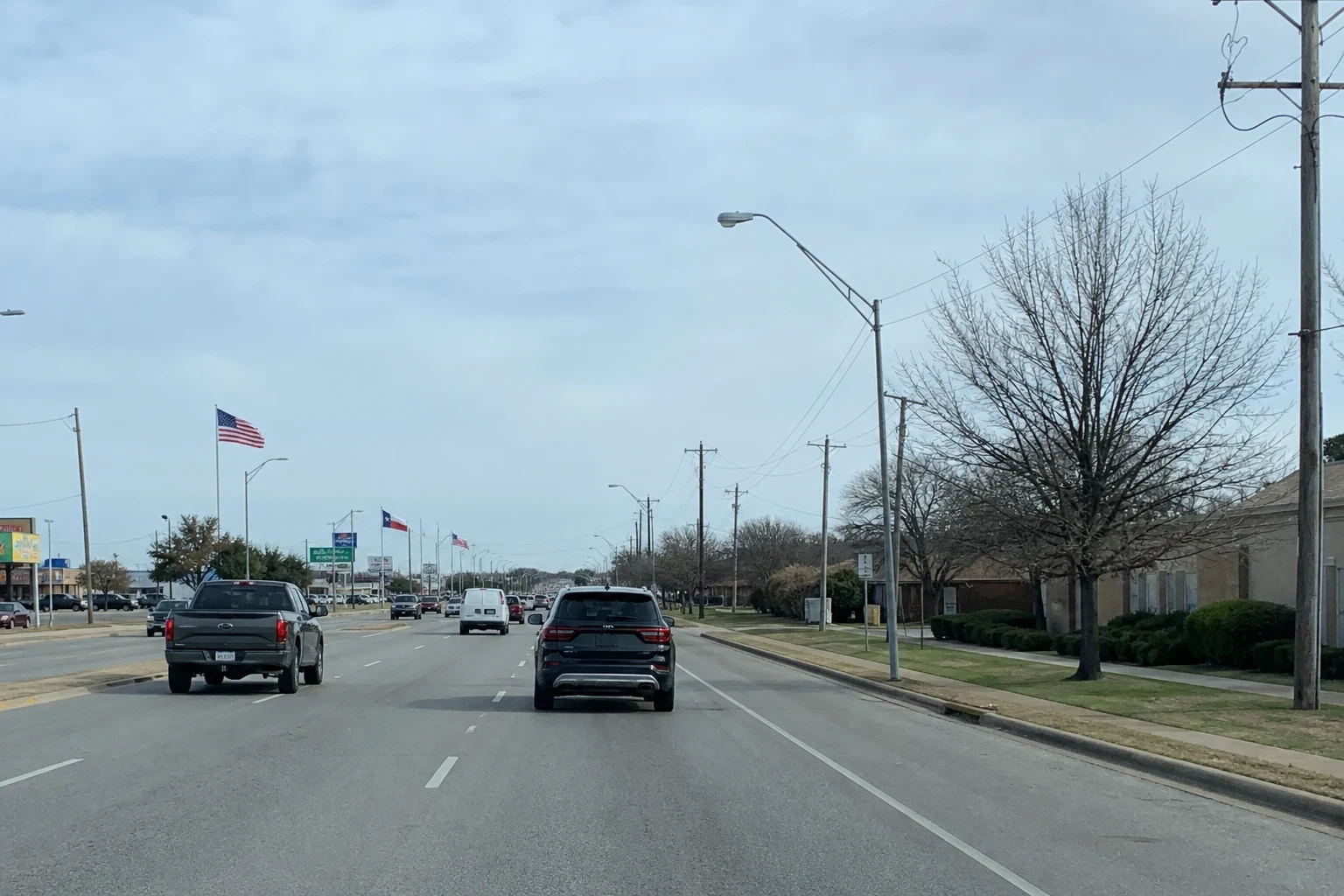 Multi-lane suburban Texas road with a pickup, SUV and van, bare winter trees, utility poles, low brick buildings and U.S. and Texas flags under an overcast sky